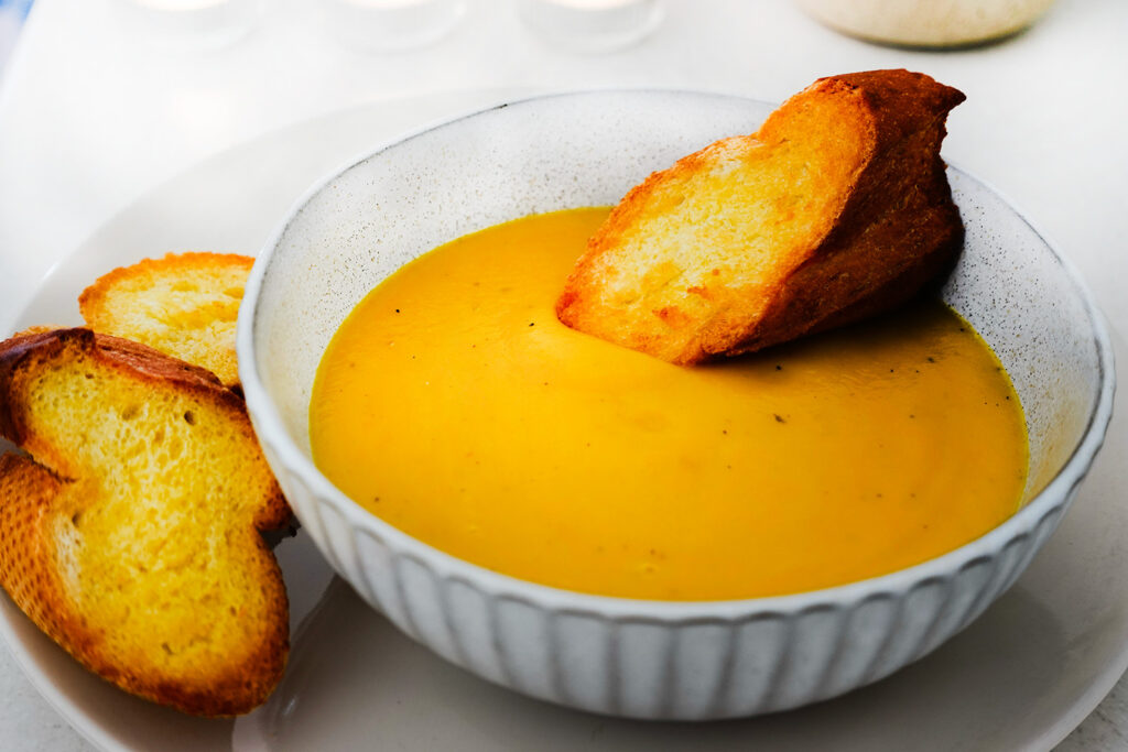 A warm bowl of creamy pumpkin soup served with a slice of golden, crusty bread on the side. The soup has a smooth texture and vibrant orange color.